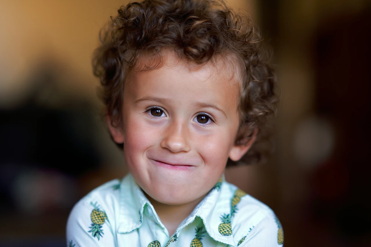 Young boy with curly hair and a slight smile, wearing a pineapple-print shirt against a softly blurred warm background.