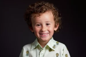 Young boy with curly hair smiling in a pale green pineapple-print shirt against a dark background.