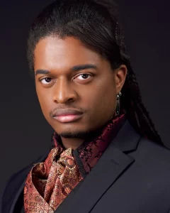 Constantine wearing a dark jacket and burgundy-and-gold patterned neckwear against a dark background.