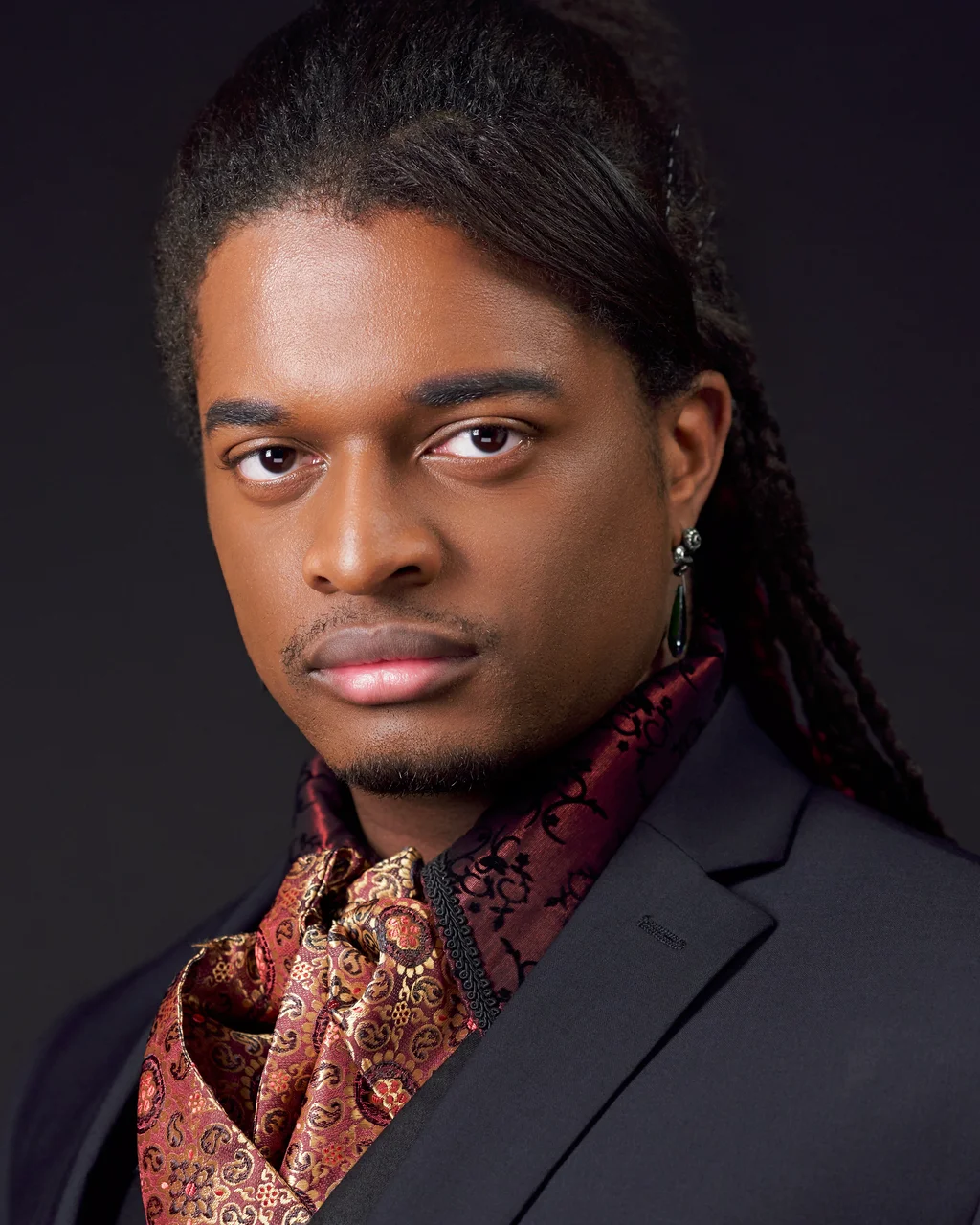 Constantine wearing a dark jacket and burgundy-and-gold patterned neckwear against a dark background.