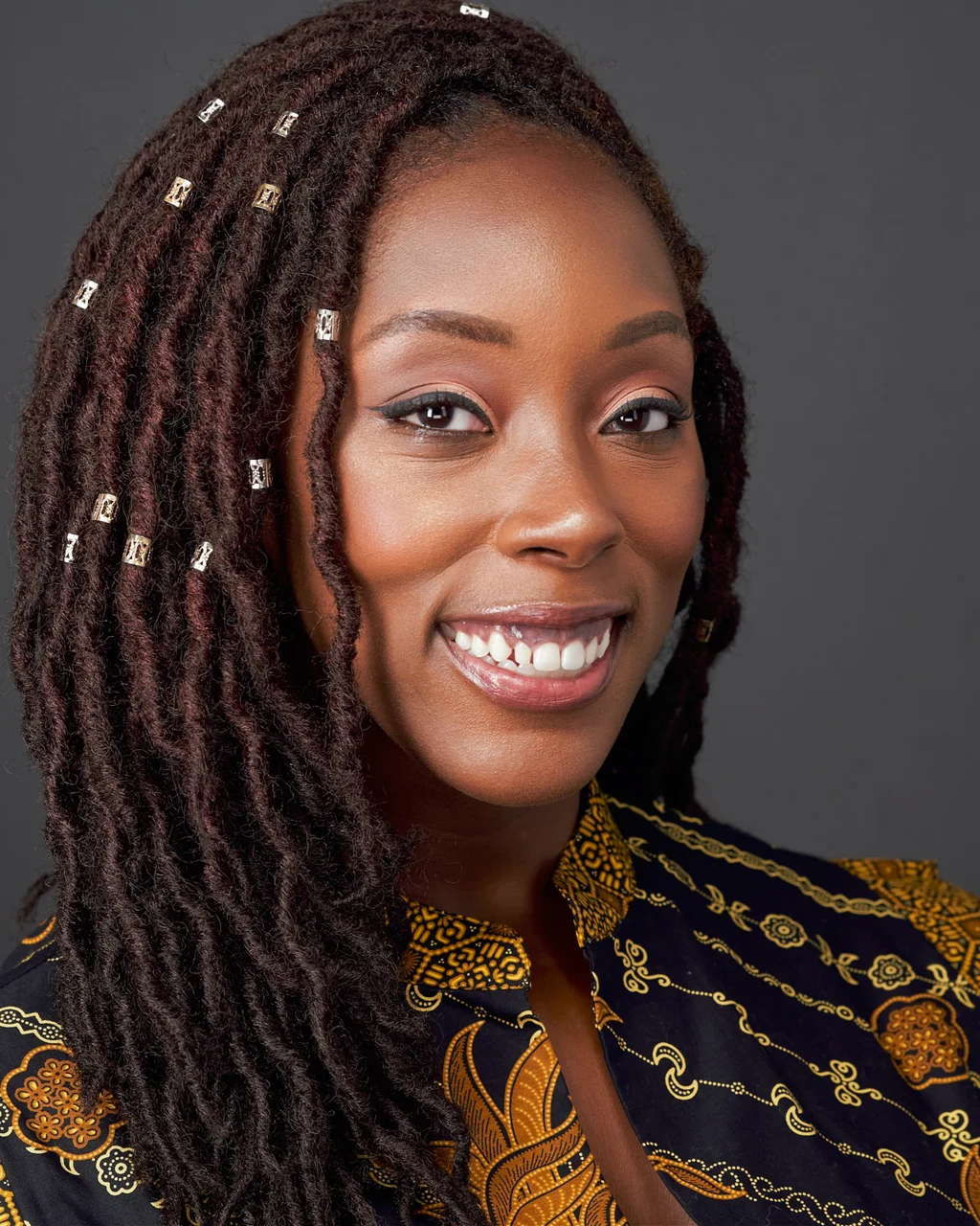 Creative professional with locs wearing a patterned jacket against a dark gray studio background.