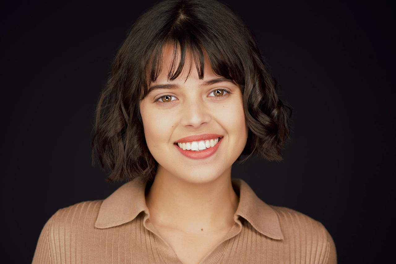 Dominique smiling with a short dark bob and bangs, wearing a tan collared top against a dark background.