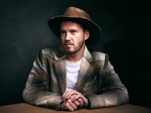 Man wearing a wide-brim hat seated at a table for a moody editorial portrait.