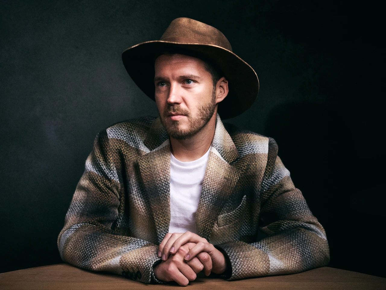 Man wearing a wide-brim hat seated at a table for a moody editorial portrait.