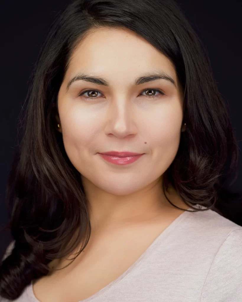 Karisma looking directly at the camera, wearing a pale mauve top against a dark background.