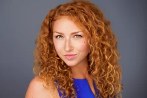 Woman with curly red hair and freckles wearing a blue sleeveless top against a gray background.
