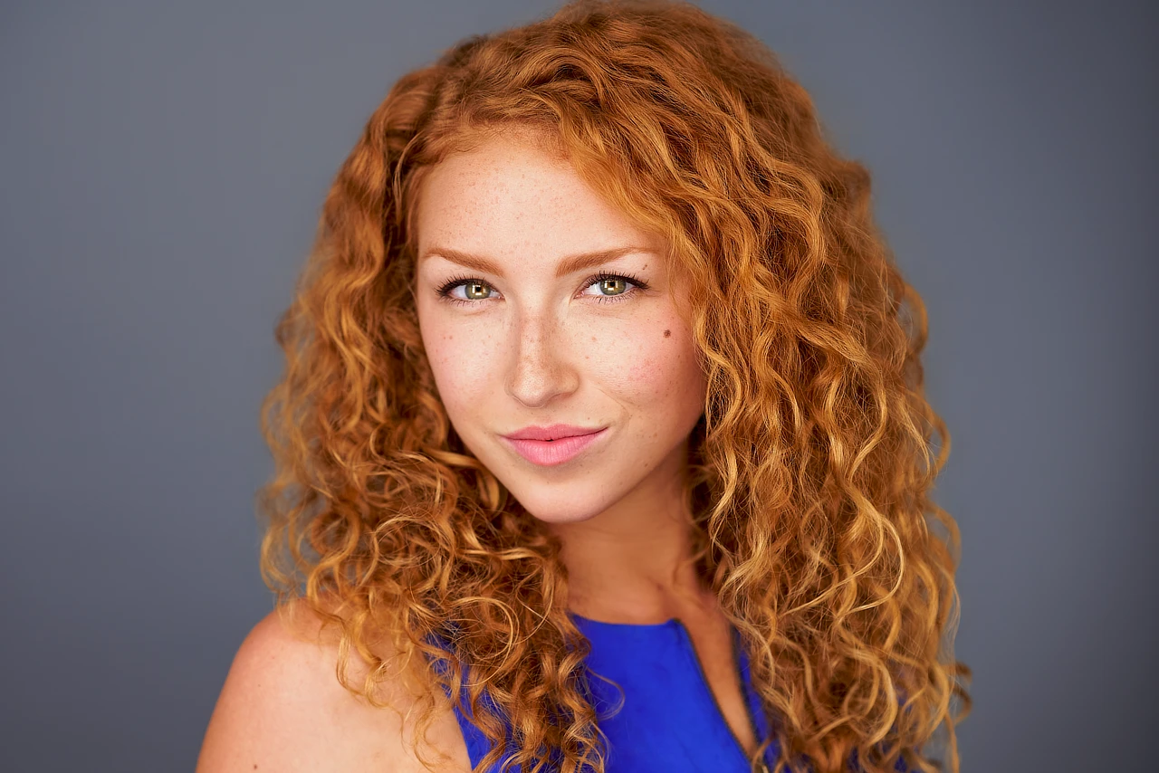 Woman with curly red hair and freckles wearing a blue sleeveless top against a gray background.