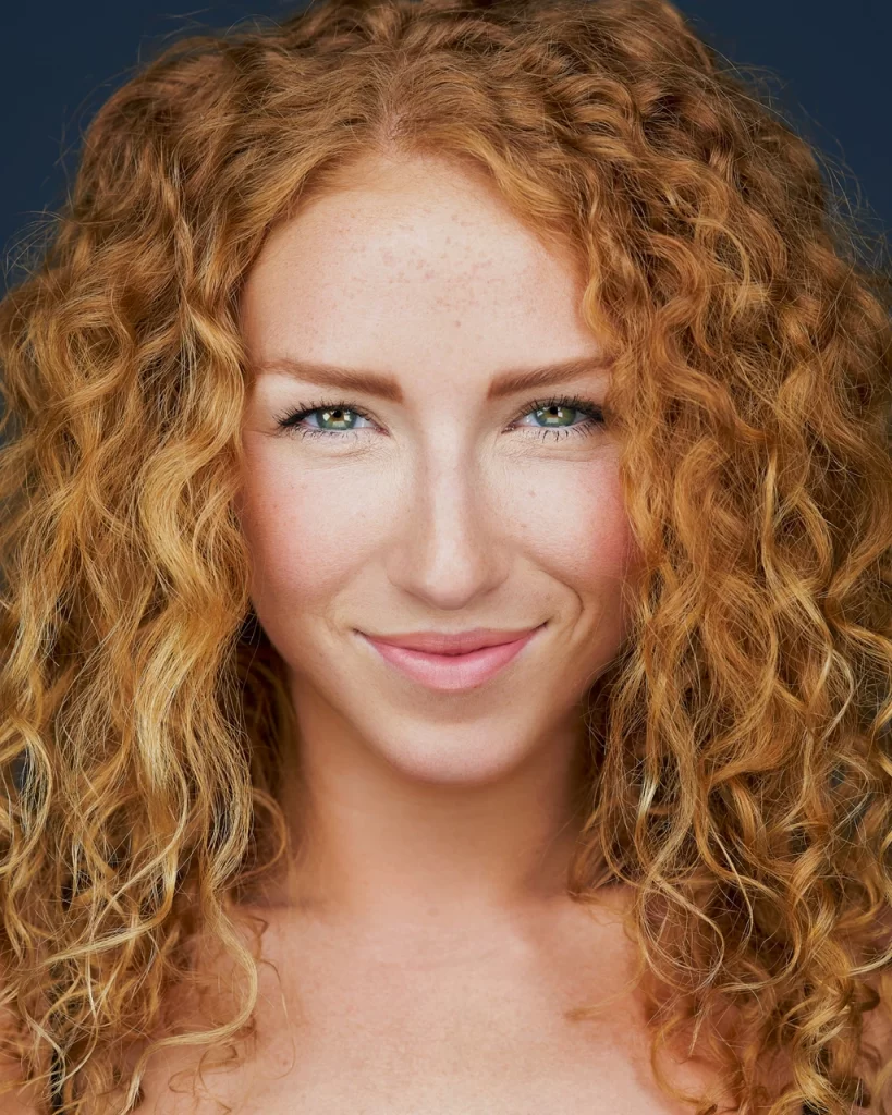Modeling headshot of red-haired woman with curly hair against a dark studio background.