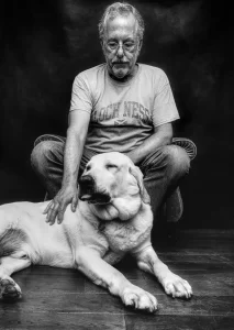 Older man seated with a large dog in a black-and-white studio portrait.