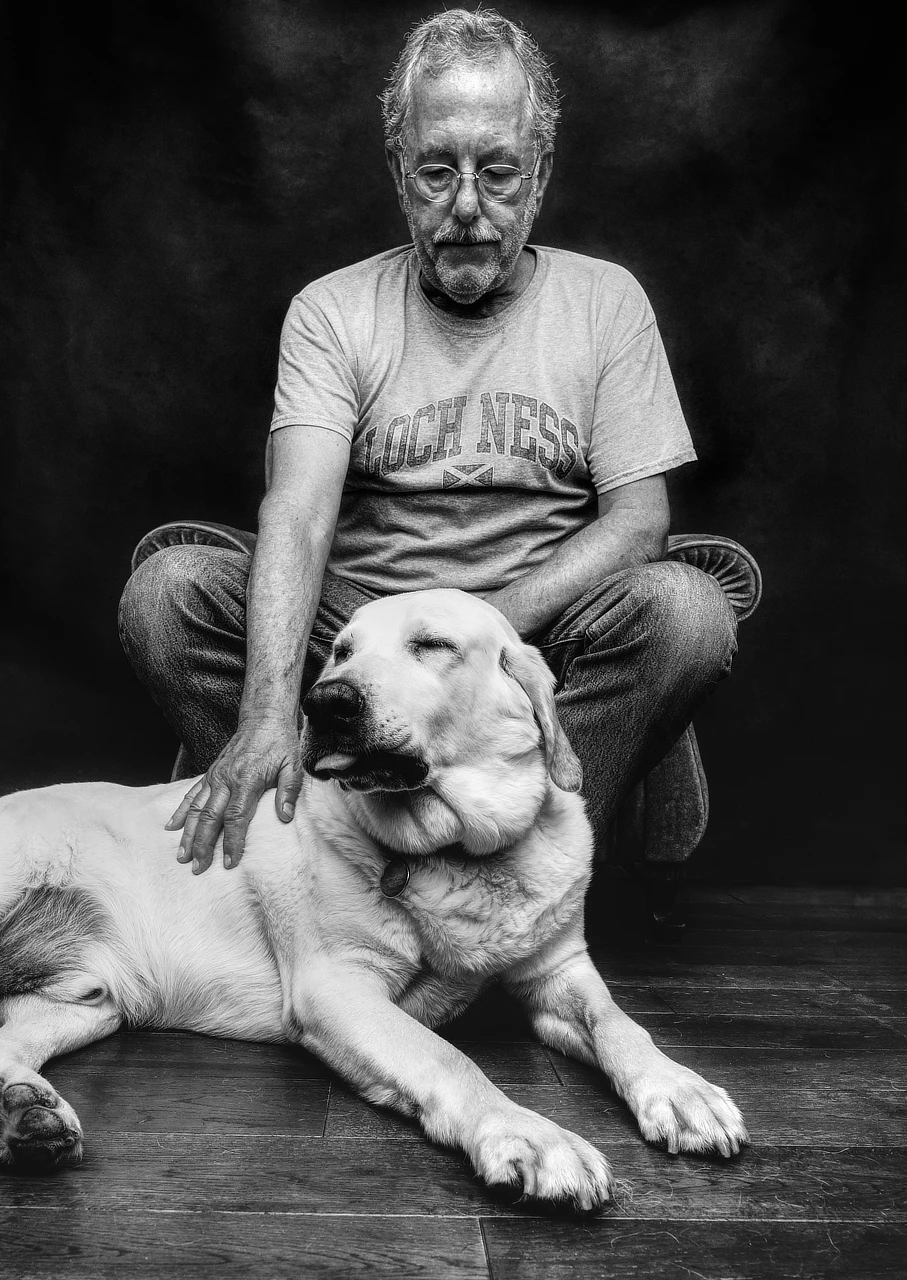 Older man seated with a large dog in a black-and-white studio portrait.