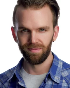 Ryan with swept-back hair and a beard wearing a blue plaid shirt against a white background.