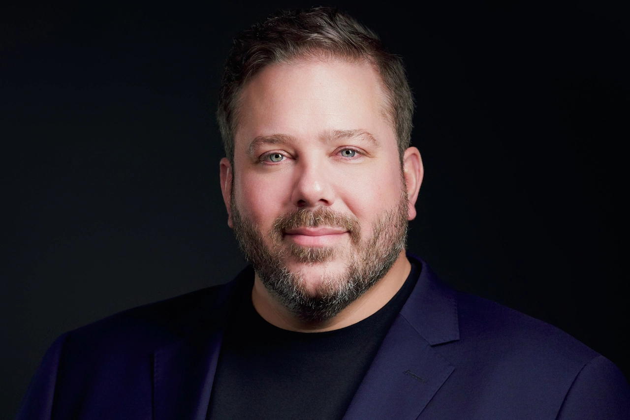 Startup founder in a dark blazer and black shirt photographed against a dark studio background.
