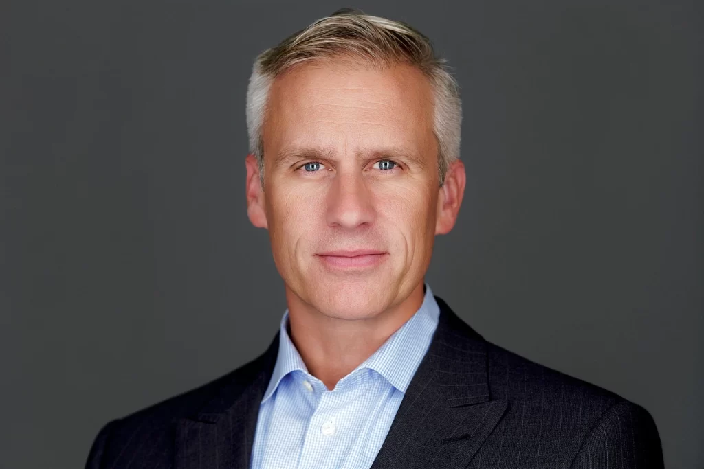Professional headshot of a finance executive in a dark suit and light blue shirt against a gray background.