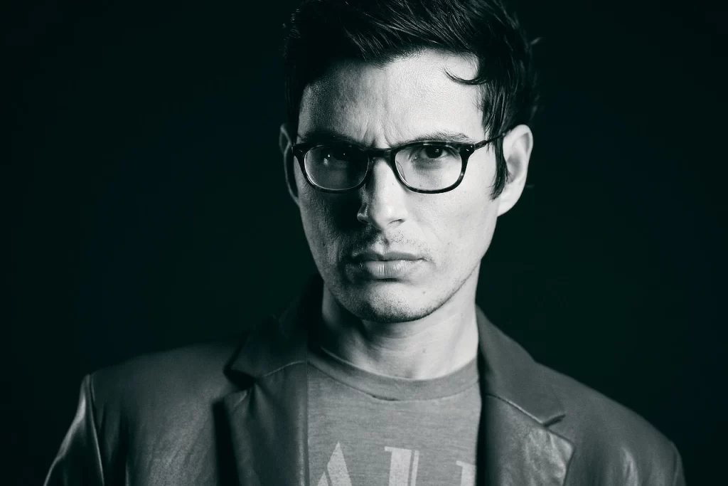 Black-and-white editorial modeling portrait of Victor wearing glasses against a dark studio background.